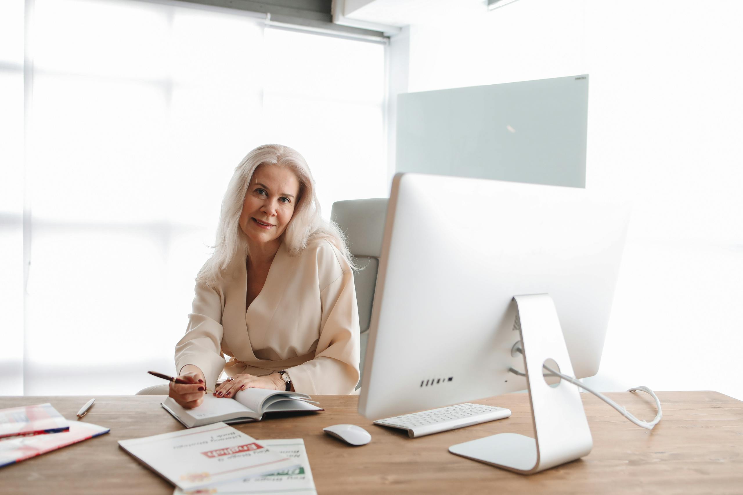 Elegant senior woman writing at office desk with computer, showcasing a professional work environment.